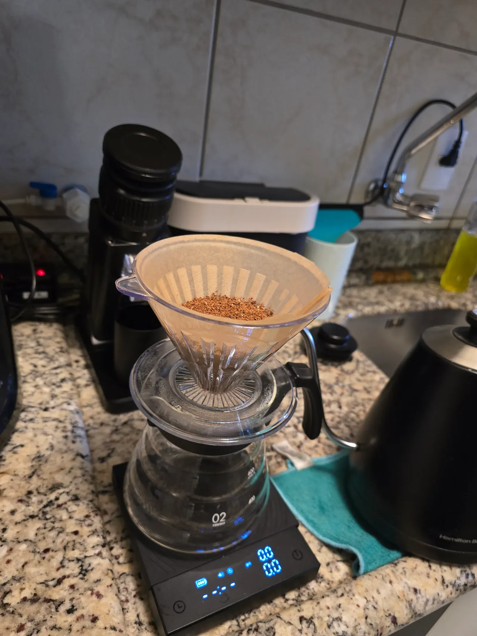 V60 dripper with a paper filter and fresh grounds on a glass server, sitting on a scale at zero, with the grinder and kettle behind