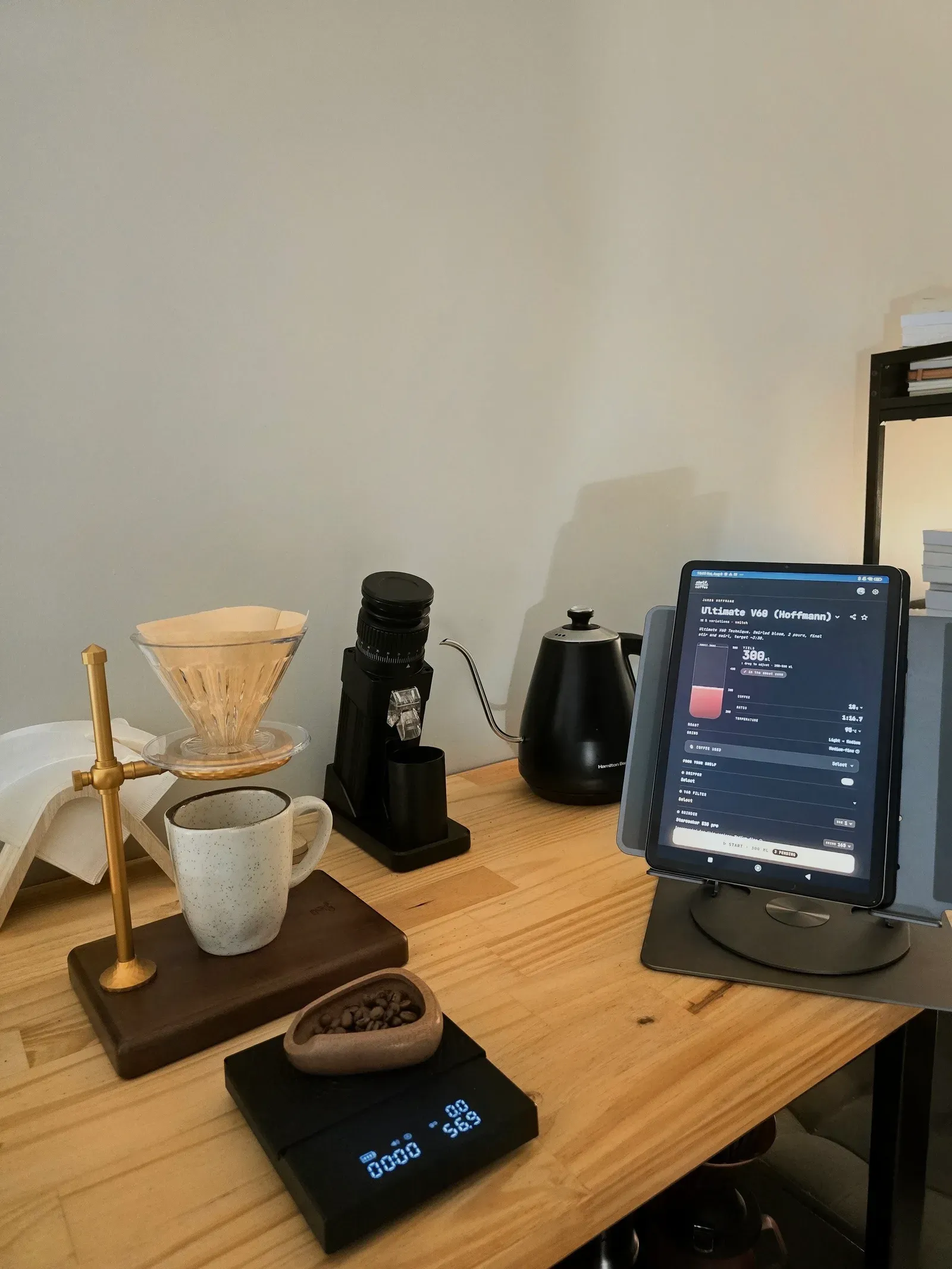 Pour-over station on a wooden desk: V60 on a brass stand over a speckled mug, 56.9 g of beans in a dish on the scale, grinder, kettle, and a tablet showing the Ultimate V60 recipe