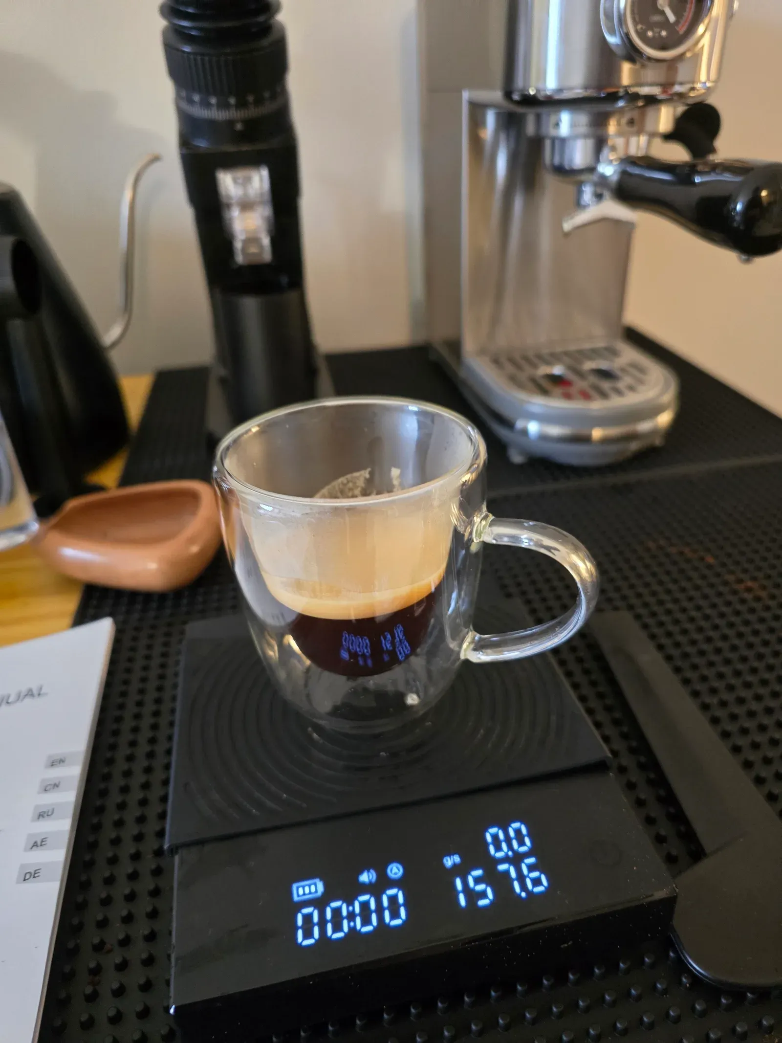 A fresh espresso in a double-walled glass cup on the scale, with the espresso machine and grinder behind it