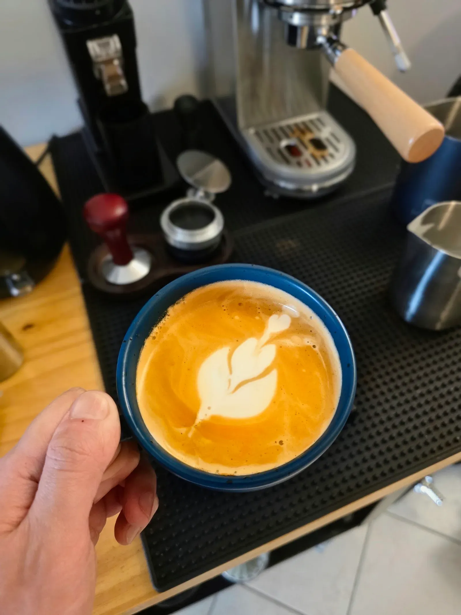 Hand holding a teal cup with a leaf-shaped tulip pour, espresso machine out of focus behind